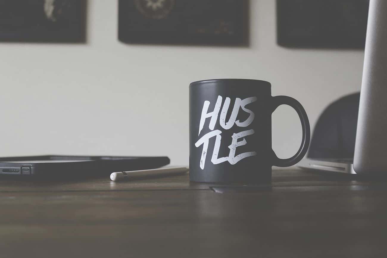 Cană personalizată neagră cu mesaj motivațional „Hustle” pe un birou modern, alături de un laptop și un stylus.